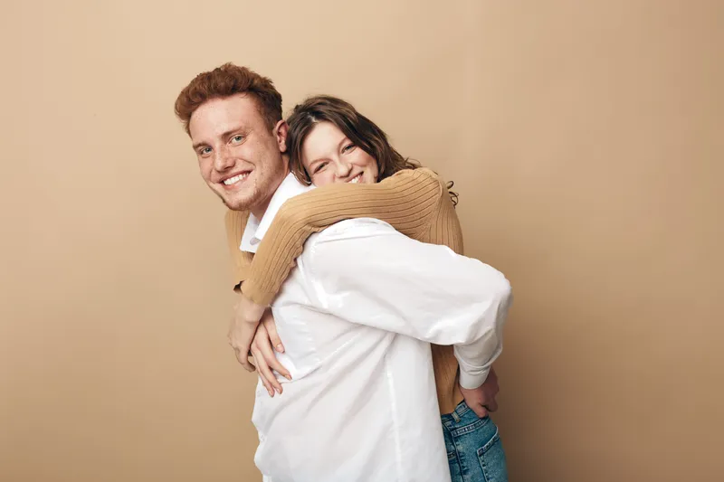Happy young couple embraces and smiles in casual clothes on beige background. The woman hugs the man from behind showing affection and joy. Concept of love, friendship, and togetherness.