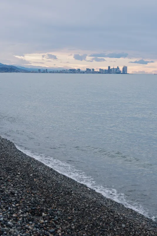 Pebble beach beside calm sea with distant city skyline on the horizon, waves touch the shore under soft sunset light across the tranquil coastal water