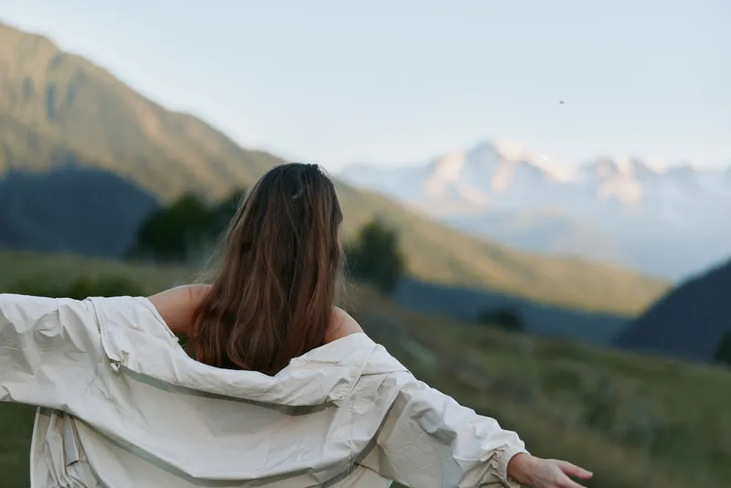 A person stands with arms wide, facing a scenic mountain landscape in a light jacket. The scene conveys freedom, serenity, and outdoor adventure in a calm natural setting.