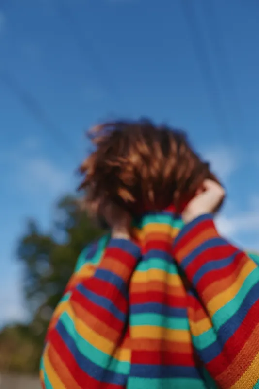 Striped sweater teen girl back view outdoor portrait under blue sky, casual outfit with short hair in playful pose adjusting hair, lifestyle travel vibe in sunny natural setting.
