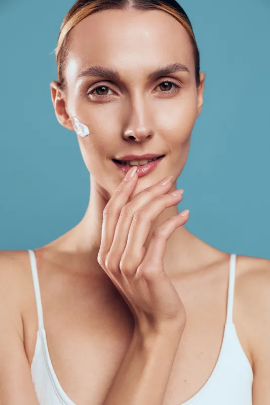 Young woman with clear skin applying white face cream on cheek, smiling gently, wearing white top, against blue background, concept of skincare, beauty, self-care, and wellness.