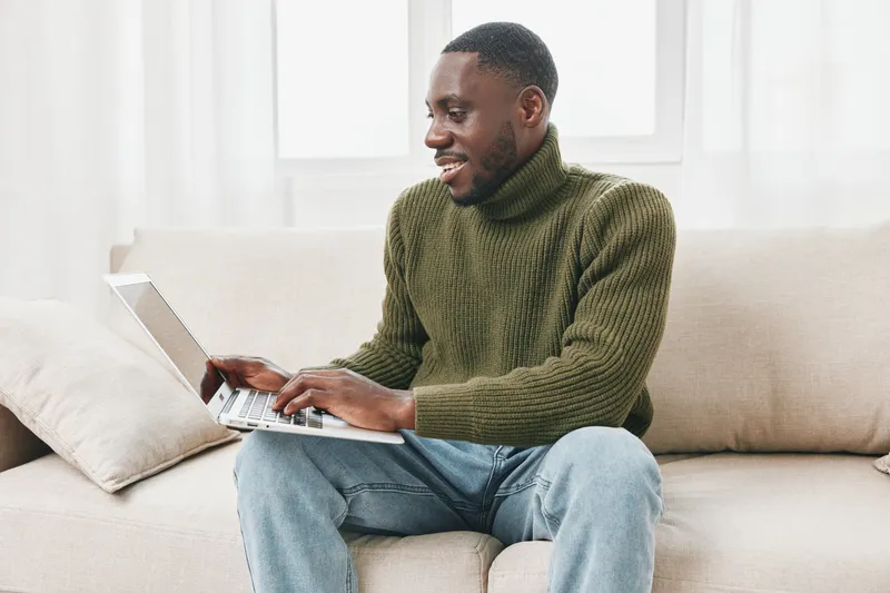 Smiling young Black man in a green turtleneck sweater using a laptop on a couch, showcasing a warm home environment with natural light