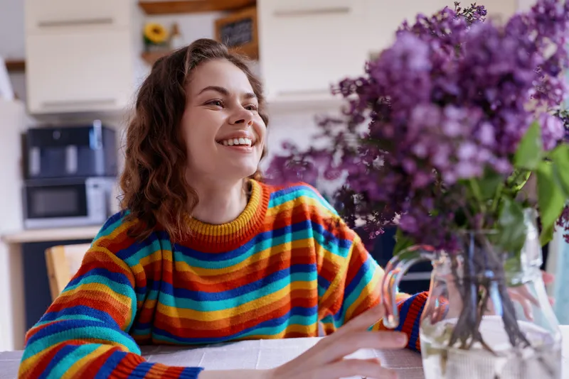 A young woman with curly hair smiles while sitting at a kitchen table in a bright striped sweater, creating a warm, inviting lifestyle moment.