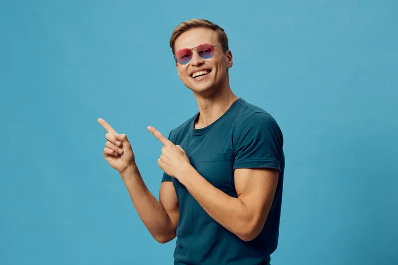 Happy young man with sunglasses smiling and pointing, casual blue T-shirt, cheerful mood, bright background, studio portrait, lifestyle concept, positive emotions, summer vibe