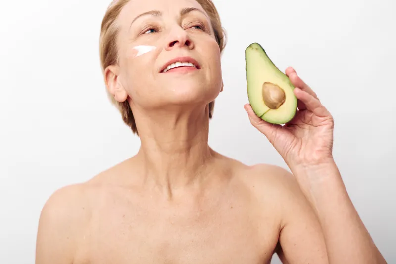 Mature woman with natural skin holds avocado half near face with cream on cheek. Concept of natural skincare, healthy aging, beauty routine, organic cosmetics, and wellness lifestyle.