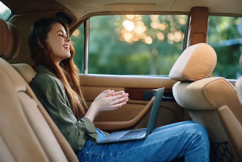 Young woman working on a laptop in the back seat of a car, enjoying a productive moment during a summer road trip, showcasing a relaxed and happy atmosphere.