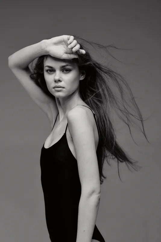 Black and white portrait of a young woman with flowing hair wearing a black tank top, expressing calm and confidence against a plain background.
