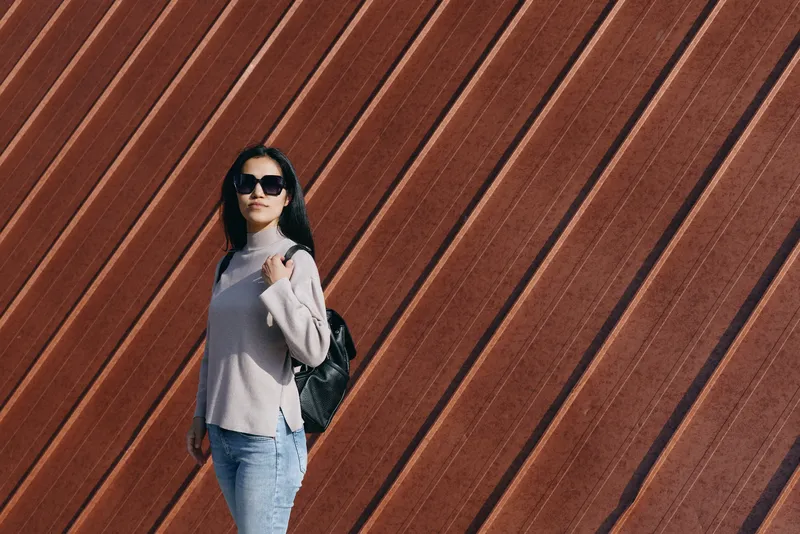Woman with backpack and sunglasses standing against a diagonal wooden wall, casual fashion on an urban outdoor street, confident pose.