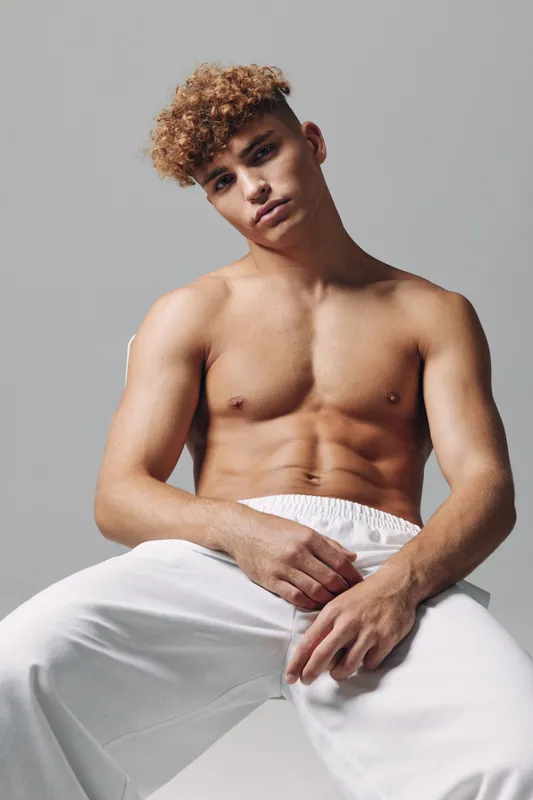 Young man with curly hair posing shirtless in white pants against neutral background. Muscular body, confident expression, studio portrait with soft lighting and casual style.