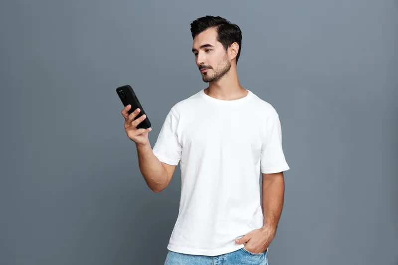 Full body thoughtful serious young caucasian man in white t-shirt looking at mobile phone stands against plain gray background studio portrait. Technology lifestyle concept