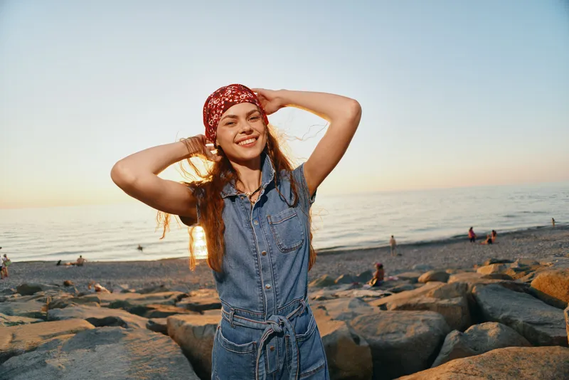 smiling woman wearing a red bandana and denim outfit enjoys a sunny beach sunset with rocks and calm sea waves in warm golden light