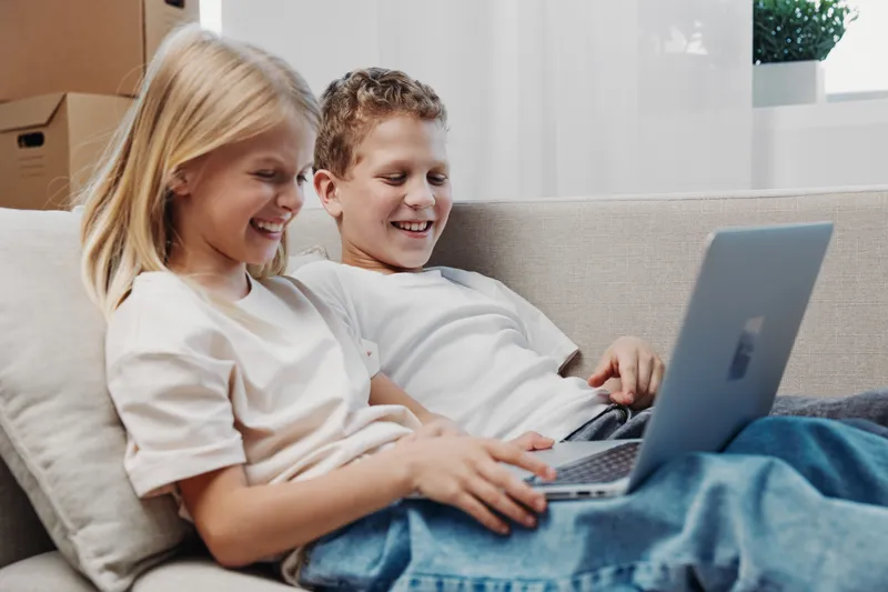 Two happy children sit on a couch using a laptop together in a bright living room. Concept for friendship, technology, and childhood leisure moments.