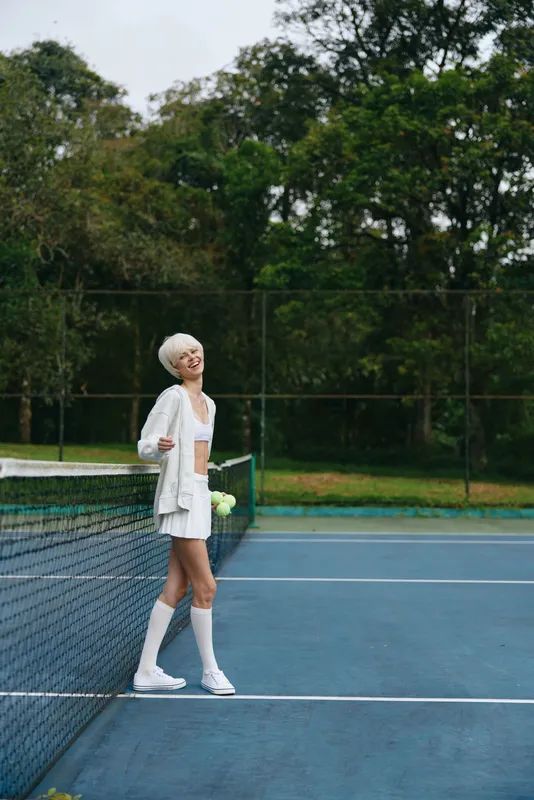 Young female athlete in tennis attire smiling confidently on the court surrounded by greenery, showcasing a vibrant yet relaxed atmosphere in sports fashion