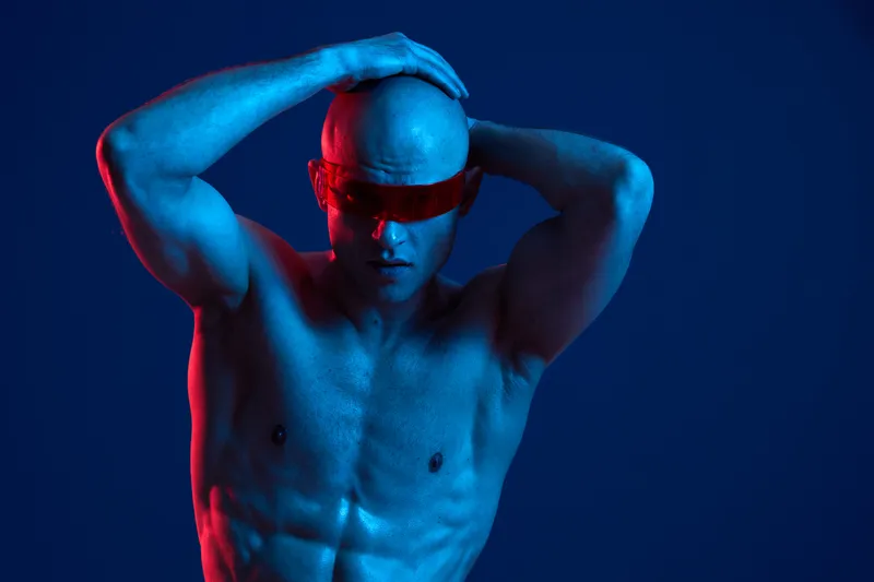 Muscular man posing dramatically with vibrant blue and red lighting, showcasing strength and style in an artistic black and white portrait.
