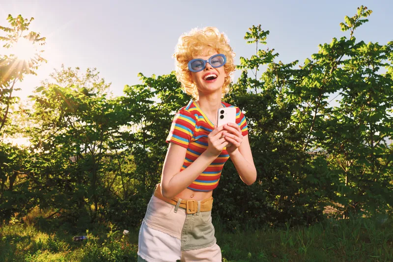 A joyful woman outdoors in bright sunlight wears a striped shirt and retro sunglasses, holding a phone and laughing. A candid lifestyle portrait capturing playful vintage charm and carefree summer