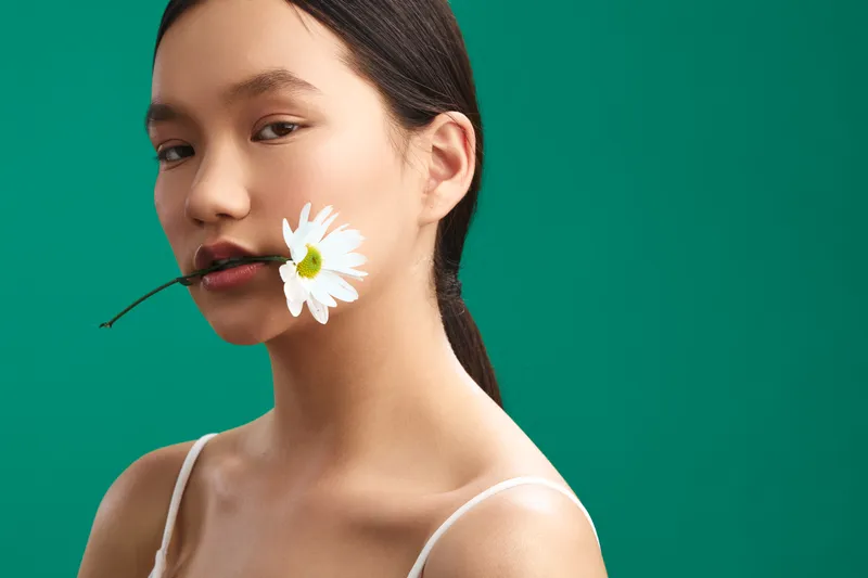 Asian woman portrait with a white daisy held near her lips, calm expression, minimal makeup, studio lighting, and a vivid green backdrop creating an artistic, fresh mood.