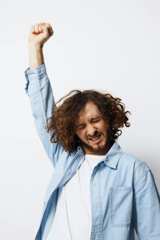 Young man with curly hair celebrating joyfully, wearing a light blue shirt against a white background, expressing excitement and positivity, suitable for motivational themes
