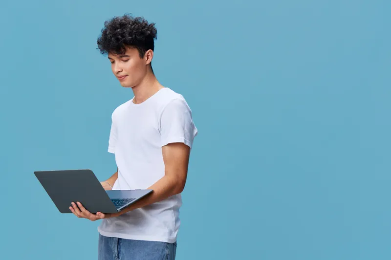 young man using laptop computer wearing casual white t-shirt standing on blue background looking focused and engaged in technology