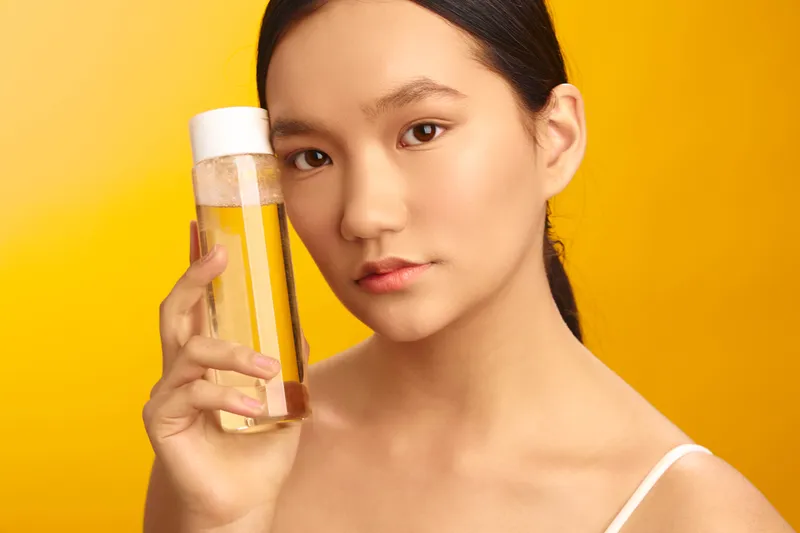 asian woman is photographed in a bright studio, holding a bottle of liquid against a yellow background. her calm expression and soft smile create a fresh, confident mood and a clean, modern aesthetic.
