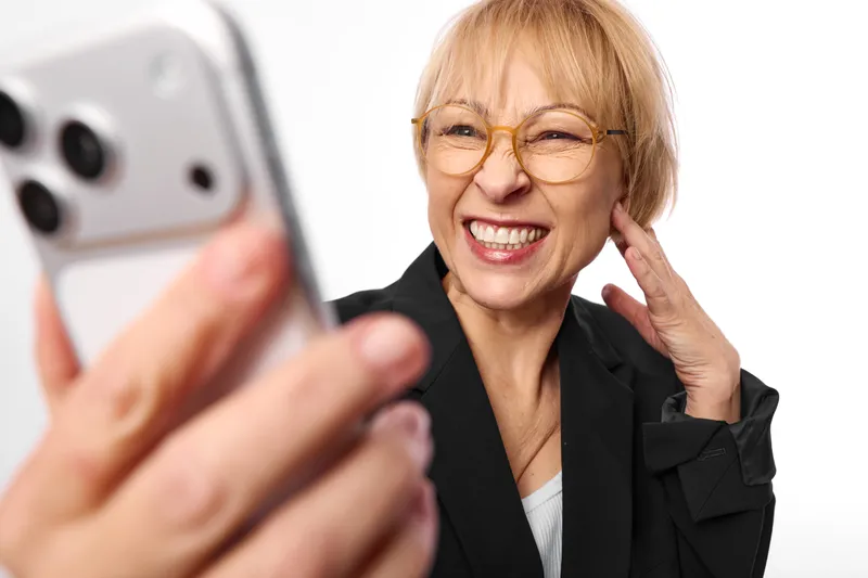 Smiling woman in business attire with glasses, holding phone near ear, blurred smartphone in foreground, warm indoor light, friendly call, white background, positive mood, portrait