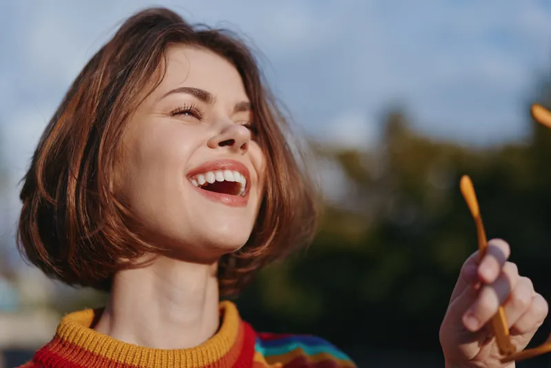 Woman smile portrait with sunglasses in hand outdoors, joyful and happy young woman with short hair wearing colorful sweater, candid sunlight and relaxed expression.