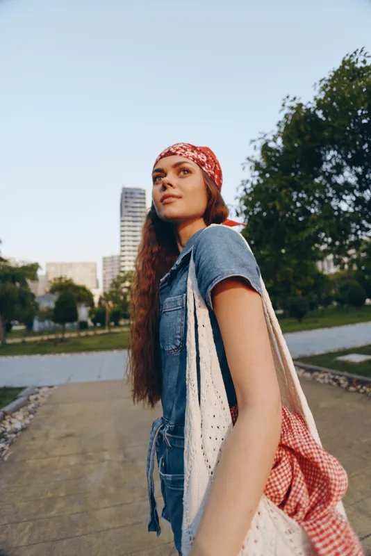 Lifestyle woman in modern boho-western denim street style with a red bandana, carrying a bag, outdoors in urban park, film color effect enhancing natural light and mood.