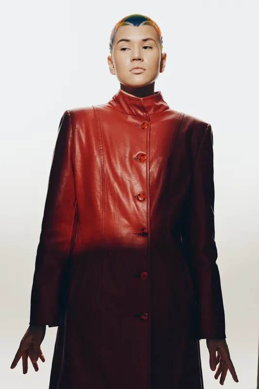 Woman with short colorful hair wearing a long red leather coat, posing confidently against a bright white background with strong lighting and shadows.