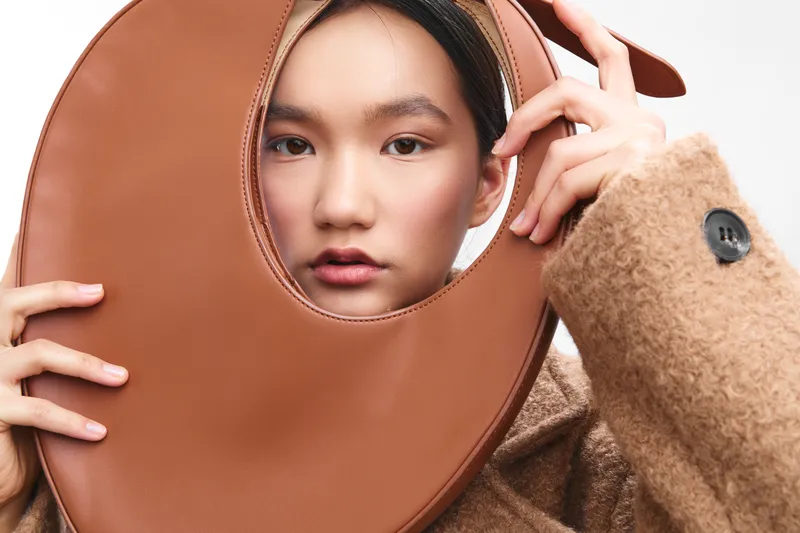 asian woman gazes softly at the camera while holding a large circular prop near her face, a fashion portrait captured in warm tones with a calm, intimate mood.