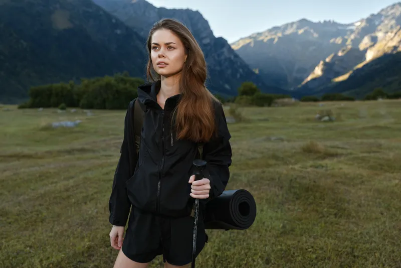 A young woman hiker stands in a wide mountain meadow, wearing a backpack and outdoor gear. She looks ahead with calm focus as rugged peaks rise behind her, inviting exploration and adventure.