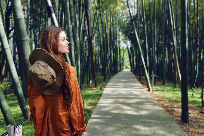Woman in orange coat with hat and smile stands on a bamboo path in a serene forest, portrait side view suggesting travel and peaceful walking amid tall green stalks and dappled sunlight