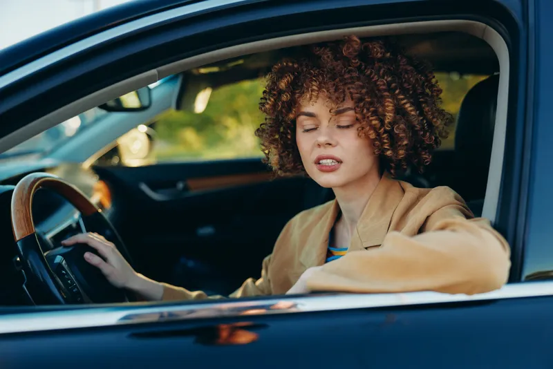A woman with curly hair sits behind the wheel of a blue car in a casual driving pose, wearing a tan jacket. Retro film color adds warmth and nostalgia to this modern lifestyle moment.
