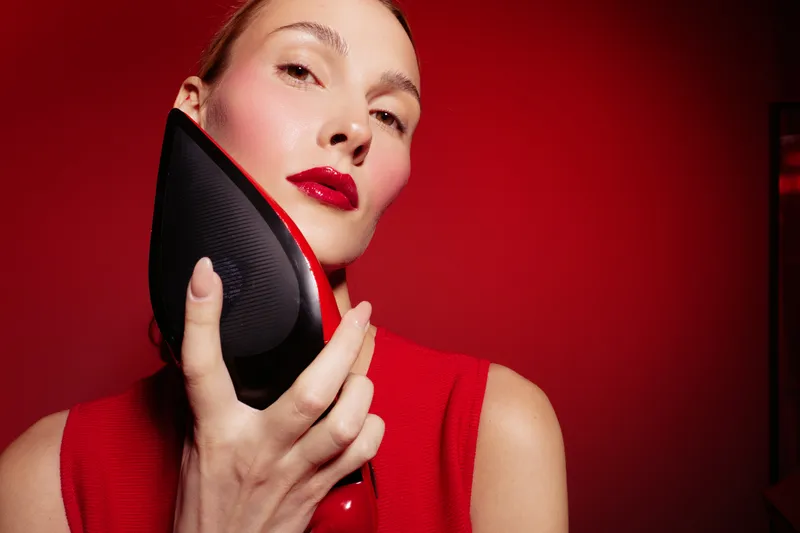 Woman with red lipstick and red top holds black and red high heel shoe close to her face against red background. Concept of fashion, style, beauty, and elegance.