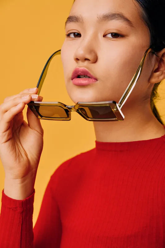Woman with sunglasses and red sweater posing on yellow background. Fashionable female model with natural makeup holding glasses for style portrait with confident expression closeup.