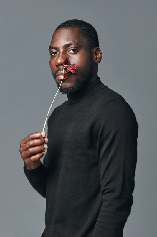 African man with dark skin wearing black turtleneck holding dried flower near face in creative conceptual visual style studio portrait with gray background and thoughtful expression.