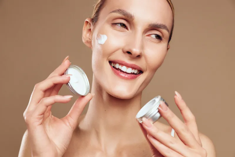 Smiling woman applies moisturizing cream on face holding open jar with beige background skin care concept natural beauty healthy lifestyle close up portrait.