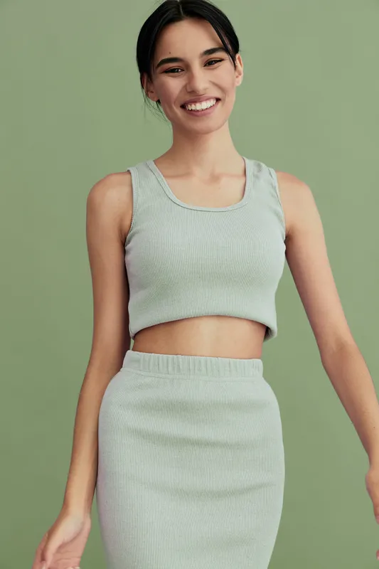 smiling woman in casual light gray knit crop top and skirt stands against green background, showing natural beauty, relaxed pose, and youthful confidence