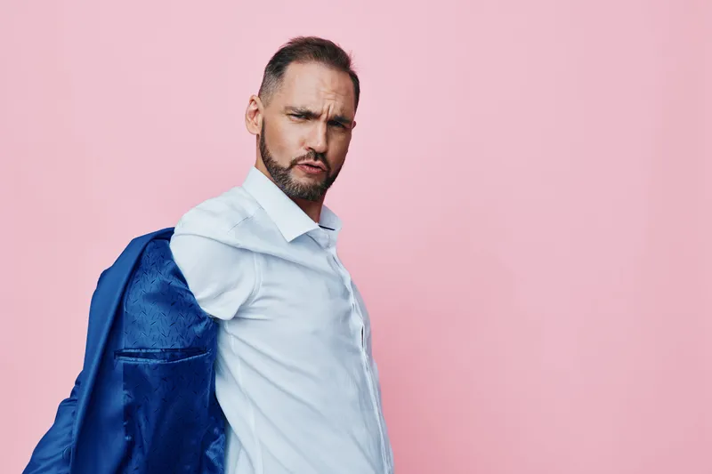 A confident male business professional stands against a pink background, exuding focus and determination with a jacket draped over the shoulder, ready for action.