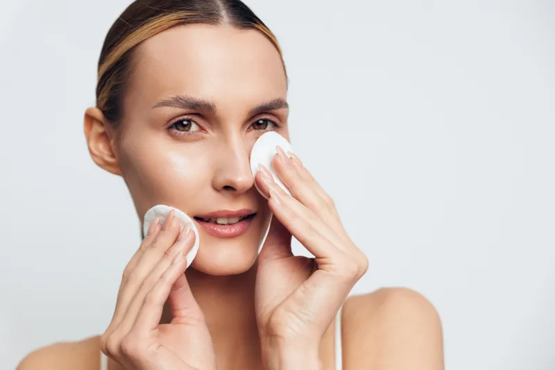 A young woman gently removes makeup using cotton pads on her clean face against a plain white background. Concept for skincare, beauty routine, and facial cleansing at home.