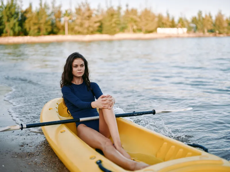 A woman sits in a yellow kayak on a calm shoreline, wearing a blue long sleeve swimsuit, gazing into the distance as gentle waves lap the shore.