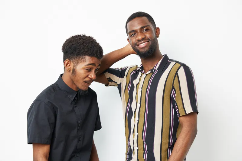 Two smiling African American men pose together, showcasing their stylish outfits and joyful expressions against a clean backdrop, highlighting friendship and camaraderie.