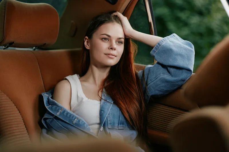 Relaxed young woman with long hair in denim jacket sitting in a car, contemplating thoughtfully, showcasing a peaceful and natural ambiance.