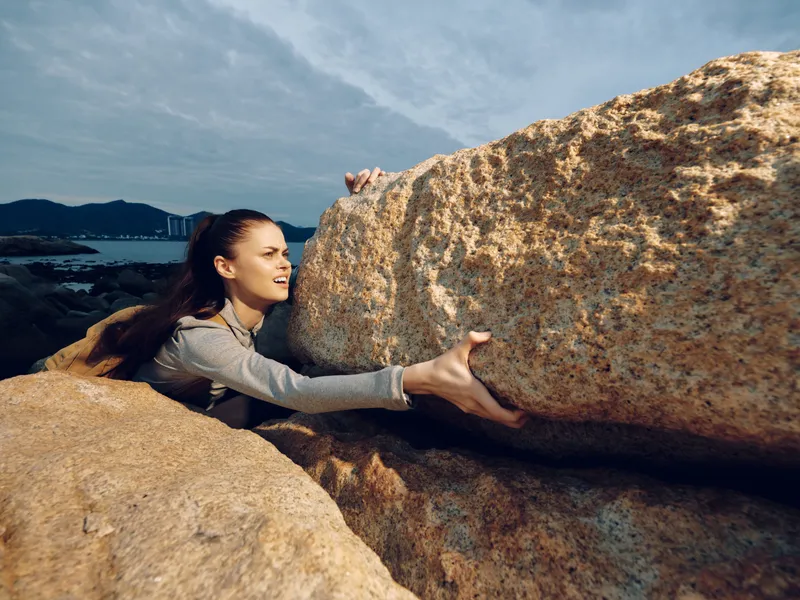 A determined young woman in activewear, climbing rock formations during an overcast day, symbolizing strength and resilience in challenging environments
