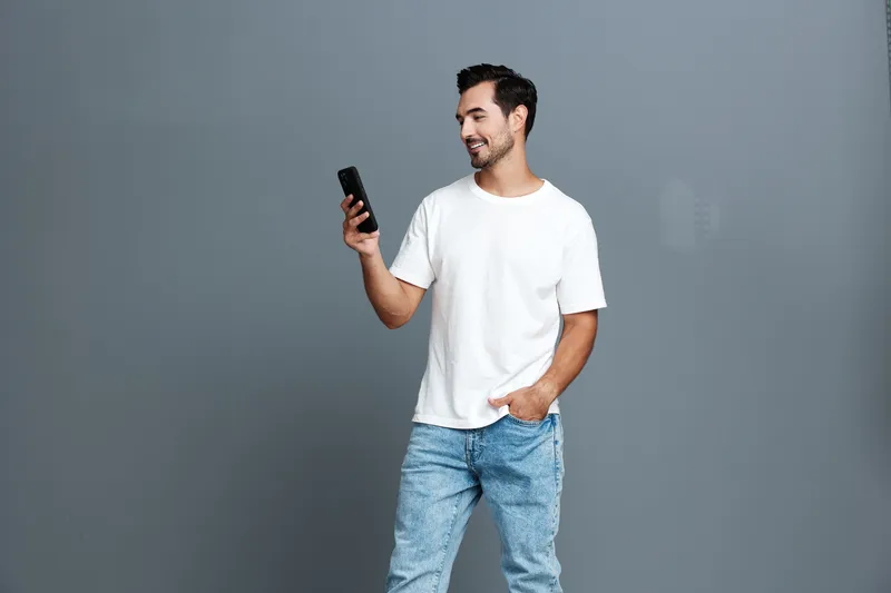 Smiling young man in casual white t-shirt stands holding smartphone, looking at screen with joy on his face, isolated on grey background. Lifestyle concept.
