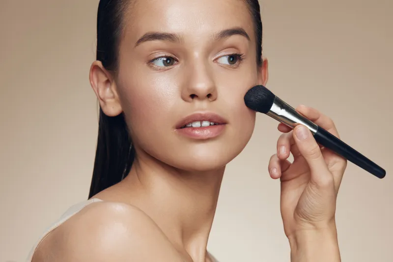 Portrait of a young woman applying makeup with a brush, showcasing a natural beauty look and glowing skin, perfect for beauty and skincare concepts