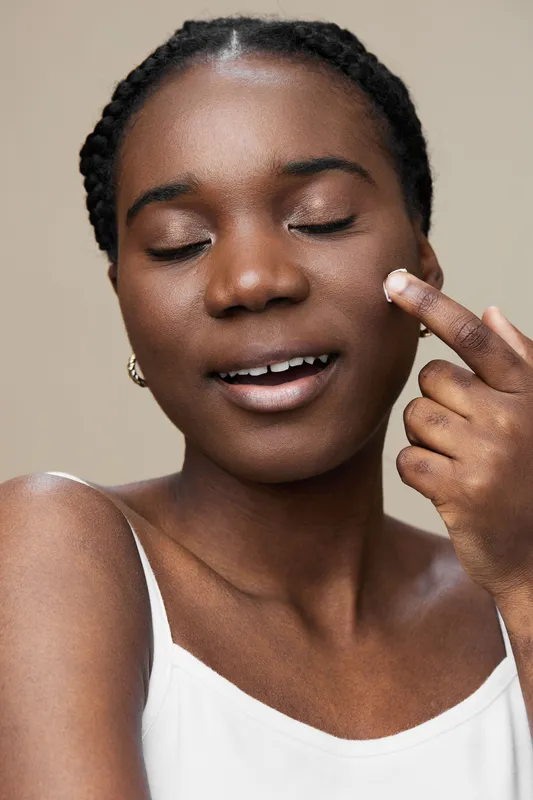 A young African woman happily applying skincare on her face, showcasing a healthy glow against a neutral background, perfect for beauty and skincare concepts