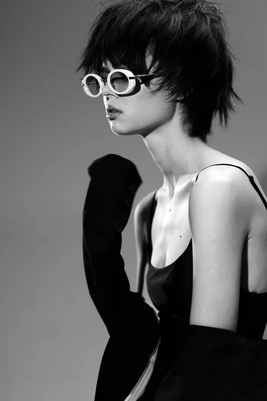 Black and white portrait of a woman with short hair wearing stylish sunglasses and a sleeveless top in a minimalist studio setting.