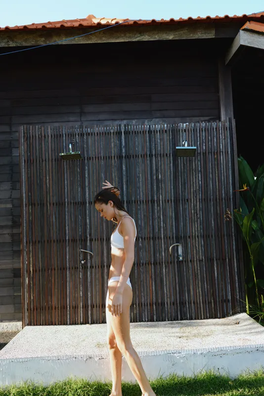 Young woman in white bikini walks barefoot on grass near outdoor wooden shower with tropical plants around, enjoying summer lifestyle and warm sunny day outside.