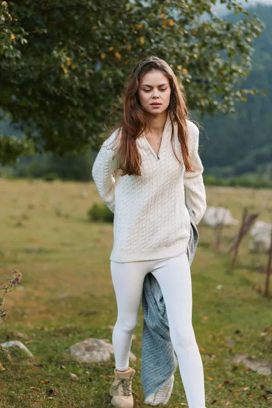 A young woman in a cream knit sweater walks across a grassy field with hills in the background, looking thoughtful.