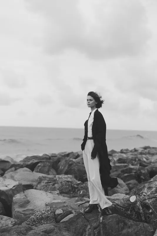 Woman stands on rugged coastal rocks, wearing a long coat and flowing dress, monochrome scene capturing wind, sea spray, and timeless fashion by the shore.