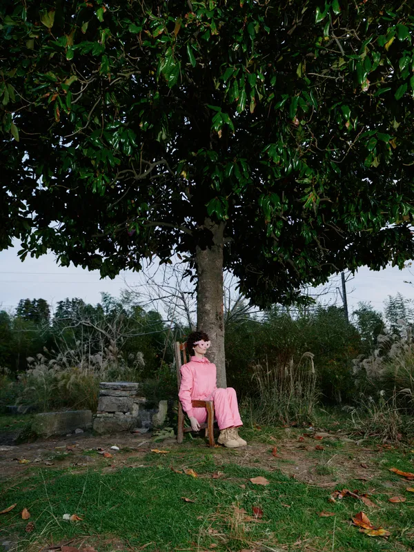 Woman in pink outfit sits under a large tree in a calm outdoor setting, posing beside a grassy area with foliage and distant shrubs, conveying quiet mood and leisure